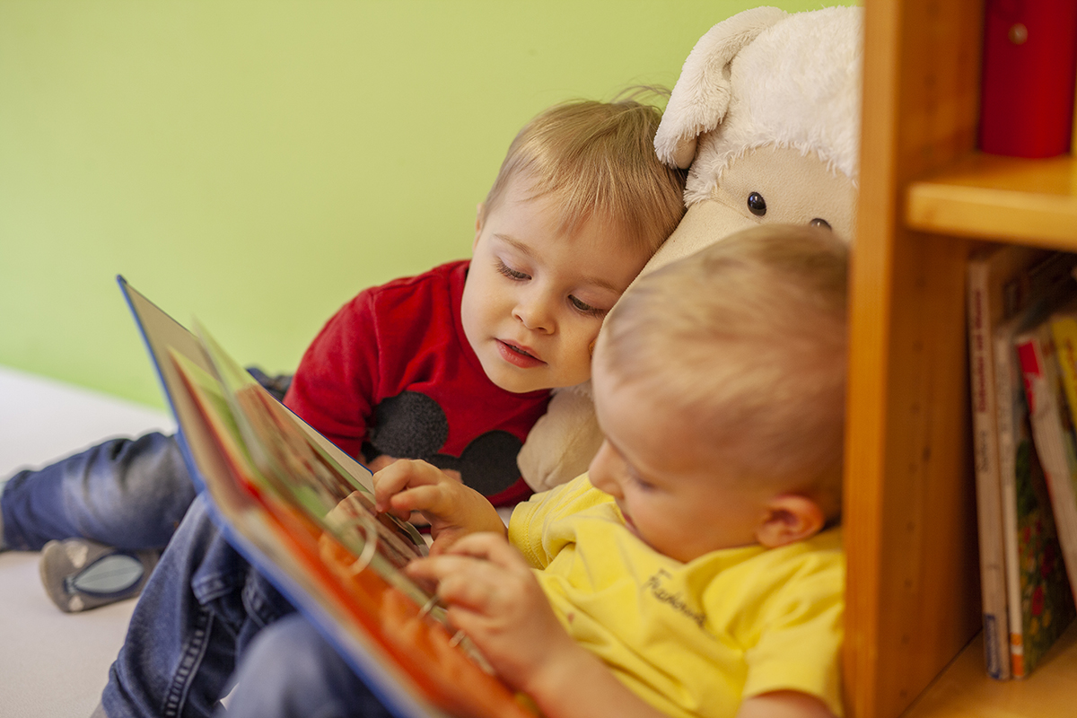 Zwei Kleinkinder schauen sich ein Buch gemeinsam an, während sie im Kindernest liebevoll von der Tagesmutter betreut werden.
